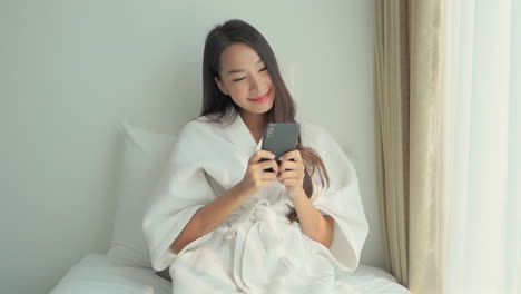 Smiling-young-asian-lady-scrolling-on-her-phone-on-bed-in-a-bathrobe