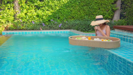 Woman-sitting-in-a-swimming-pool-with-floating-tray-of-breakfast-smelling-coffee