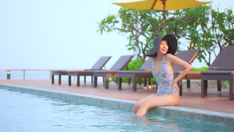Asian-female-wearing-a-swimwear-and-floppy-hat,-sitting-at-the-swimming-pool-with-her-feet-submerged-in-the-water-in-a-luxurious-hotel