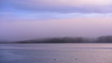 Fog-over-sea-and-seaside-town-at-sunrise-with-calm-water-and-colourful-sky