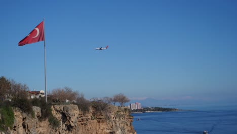 Full-shot,-turkey-flag-on-the-flagpole,-besides-the-cliff-of-Antalya,-Aeroplane-descending-in-the-background