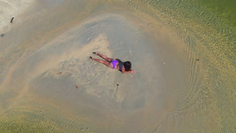 Aerial-view-of-a-young-woman-laying-on-a-sandbar-in-a-river-wearing-only-a-sexy-purple-bikini