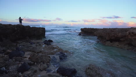Einsamer-Mann-Beim-Angeln,-Während-Die-Wellen-Bei-Sonnenuntergang-In-Einer-Kleinen-Felsigen-Bucht-Am-Mokuleia-Rock-Beach---Oahu-Hawaii-Gegen-Das-Ufer-Schlagen