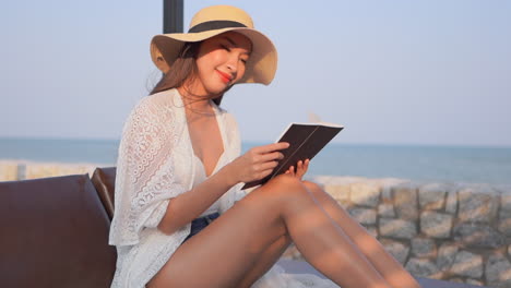 Asian-tourist-reading-a-book-and-wearing-a-sun-hat-while-sitting-near-the-ocean-with-wind-softly-blowing-her-hair