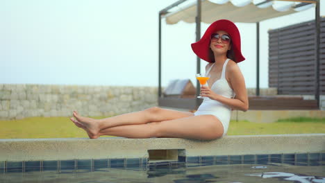 Sexy-female-tourist-sitting-near-a-swimming-pool,-wearing-a-one-piece-swimwear-and-a-red-floppy-hat-enjoying-her-vacation