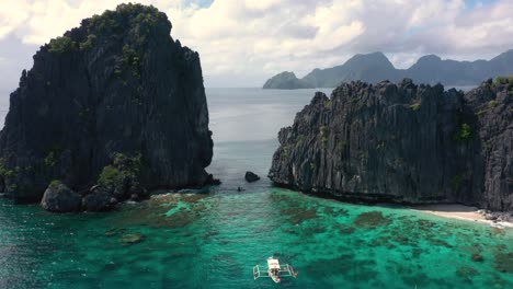 Aerial-of-huge-limestone-cliffs,-turquoise-water-and-natural-archipelago-with-drone-flying-through-rocks-and-small-boats-in-El-Nido,-Palawan,-Philippines