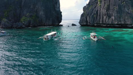 Aerial-of-huge-limestone-cliffs,-turquoise-water-and-natural-archipelago-with-drone-flying-through-rocks-and-boats-in-El-Nido,-Palawan,-Philippines