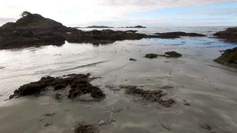 Aerial-view-from-drone-flying-low-over-wet-sand,-rocks-and-tidal-pools-toward-open-water-on-coast-of-Vancouver-Island-near-the-village-of-Tofino