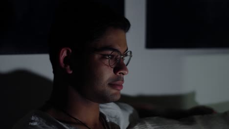 Side-View-Of-A-Young-Handsome-Mixed-Race-Indian-Man-In-Eyeglasses-Sitting-On-A-Couch,-Enjoys-Watching-Television-Alone-In-A-Dark-Living-Room