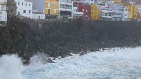 Eine-Große-Welle-Des-Atlantiks-Bricht-An-Einem-Sonnigen-Tag-Während-Eines-Sturms-In-Puerto-De-La-Cruz-Auf-Den-Kanaren-An-Einer-Felsigen-Küste,-Ferne-Bunte-Häuser,-Weitwinkelaufnahme