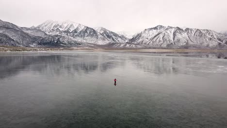 Ice-Fishing-on-Frozen-Lake-Surface-at-Crowley-Lake-in-Cold-Winter-California,-Person-on-Icy-Lake-Looking-For-Spot-to-Dig-a-Hole-For-Ice-Fishing,-Lakeside-Landscape-and-Snowy-Rocky-Mountains-in-Horizon