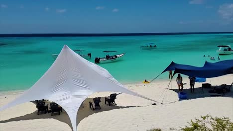 People-enjoy-beach-day,-boats-and-star-tents,-pan-left-Scenic-view-cayo-de-agua-island