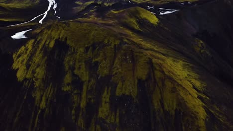 Aerial-view-over-natural-textures-and-patterns-of-the-Icelandic-highlands-dark-mountain-peaks,-covered-bright-green-grass