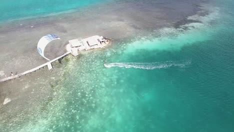 Aerial-view-Man-kiteboard-crystal-sea-water-near-iconic-palafito-stilt-house,-Los-Roques
