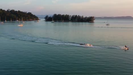 Panoramic-view-aerial-shot-capturing-water-sport-activity,-banana-boat-ride-pulling-behind-a-jet-ski-with-beautiful-landmark-of-Perdana-quay-light-house-in-the-background-at-sunset,-Langkawi-island