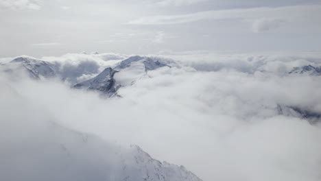 Mystic-clouds-surrounding-icy-and-snow-covered-mountains-during-sunny-day-in-Austria---Aerial-orbiting-shot