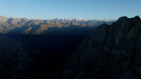 Aerial-flight-showing-dark-valley-and-bright-snowy-mountain-summits-in-background-at-sunset---dolomites-mountains-of-italy