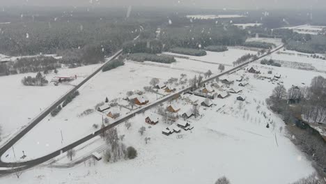 Winter-woodlands-with-small-town-and-wide-road-during-snowfall,-aerial-view
