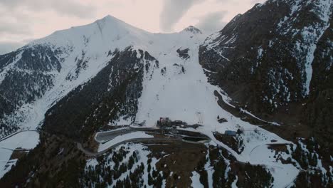 El-Dron-Vuela-Hacia-La-Cubierta-De-Nieve-Sinuosa-De-La-Carretera-De-Montaña-Vacía-Y-Las-Pistas-De-La-Estación-De-Esquí