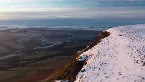 Aerial-Footage-Flying-Over-Snow-Covered-Cliff-of-the-Campsie-Fells-and-Panning-to-Reveal-Dumgoyne-Hill-with-Loch-Lomond-in-the-Distance