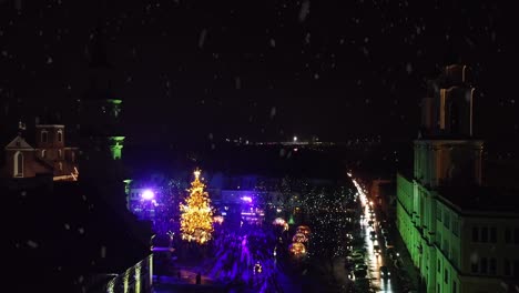 Kaunas-City-Hall-square-with-glowing-Christmas-lights-during-snowfall,-aerial