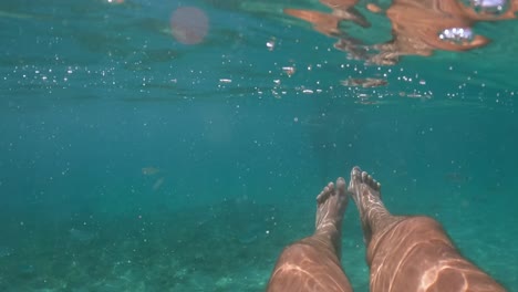 Under-water-personal-perspective-view-of-man-legs-floating-in-clear-transparent-sea-water-beneath-surface,-slow-motion