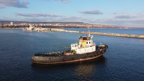 Drone-circling-industrial-boat-in-port-of-Varna,-Black-sea-with-buildings-in-the-back