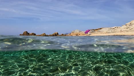 Half-underwater-point-of-view-of-Saleccia-rocky-side-of-beach-with-one-purple-sun-umbrella-open-on-shore,-Corsica-island-in-France