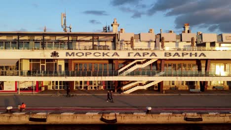People-walking-on-Port-of-Varna-pier-drone-pan-left-to-right-during-sunset-in-front-of-building