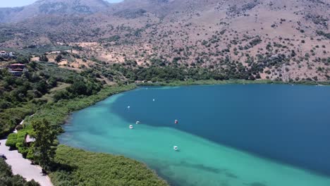 Hermoso-Lago-Kournas-En-Creta-Con-Botes-En-El-Agua-Turquesa-Que-Revela-Las-Montañas-Y-El-Cielo-Azul