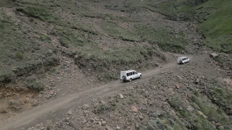 White-vehicles-drive-up-steep-gravel-mountain-pass-to-Lesotho,-Africa