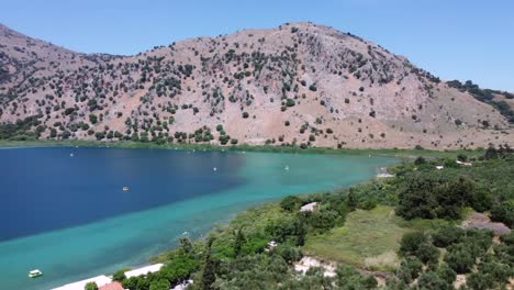 Hermoso-Lago-Kournas-En-Creta-Con-Botes-En-El-Agua-Turquesa,-Cielo-Azul-Y-Montañas-En-El-Fondo