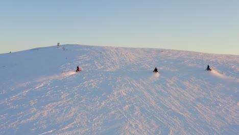 Aerial-view-snowmobile-speeding-up-snowy-sunlit-hillside-in-arctic-circle-with-group-of-friends