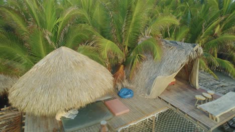 Aerial-view-of-sun-beds-on-wooden-platforms-next-to-palm-trees-by-the-ocean,-in-a-beach-club-in-Puerto-Escondido,-Mexico