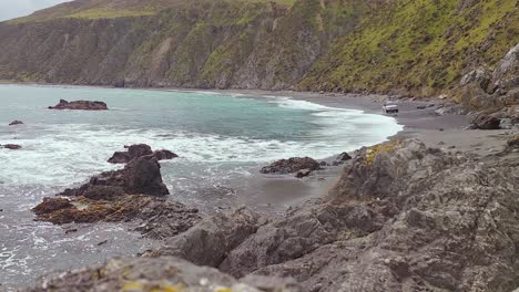 A-4WD-driving-along-a-narrow-beach-in-Cave-Bay,-Wellington,-NZ
