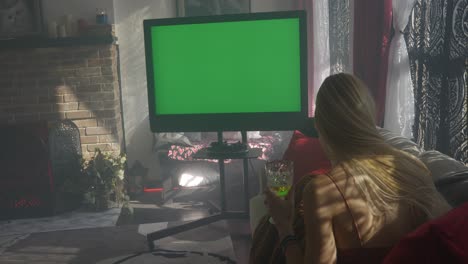 Green-screen-television-on-misty-hazy-morning-with-blonde-women-lady-girl-sitting-on-couch-cuddled-up-with-blanket-as-someone-hands-her-champagne-wine-glass-as-she-watches-the-tv