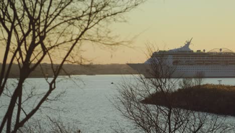 Cruise-ship-exits-port-at-sunset,-passengers-moving-on-deck-wide-shot-behind-branches