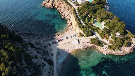 Aerial-view-of-a-beautiful-cove-with-turquoise-blue-water-surrounded-by-a-pine-forest-next-to-a-tourist-resort