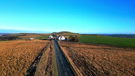 Aerial-drone-video-footage-of-a-car,-van-driving-on-a-country-road-and-passing-white-washed-buildings,-high-on-the-Pennine-Hills,-UK