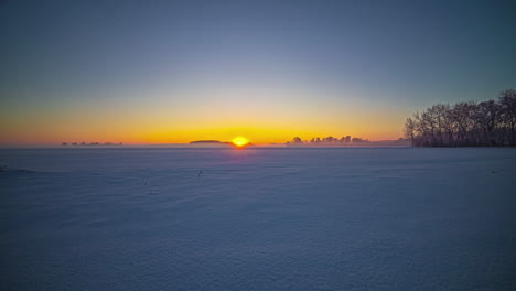 Bright,-golden-sunrise-on-a-hazy,-winter-day-with-no-clouds-across-a-field-of-snow---time-lapse