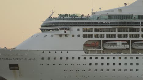 Cruise-ship-exits-port-at-sunset,-passengers-moving-on-deck-close-up-telephoto-shot