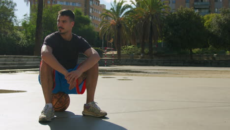 Full-shot-of-a-young-Caucasian-man-relaxing-and-resting-on-a-ball-on-a-street-basketball-court-in-Barcelona,-Catalonia,-Spain