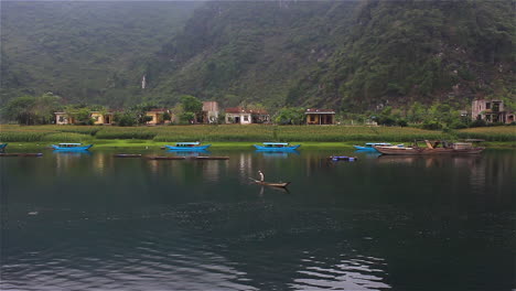 Vietnamesischer-Fischer-Auf-Dem-Fluss-In-Phong-Nha-Ke-Bang,-Nordvietnam,-Morgen-An-Einem-Nebligen-Und-Bewölkten-Tag,-Mit-Bergen,-Traditionellen-Blauen-Booten-Und-Kleinen-Häusern-Im-Hintergrund