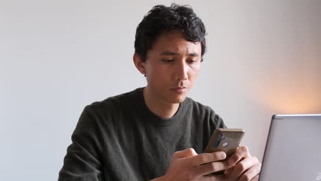 Eurasian-man-thinking-about-a-text-he's-writing-on-his-phone-while-working-from-home-on-his-computer