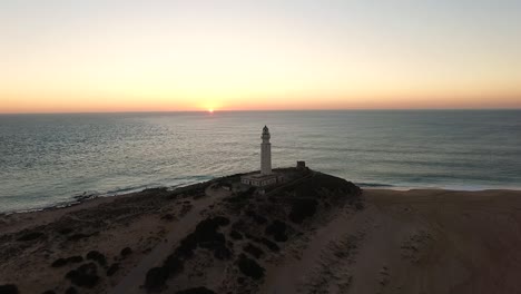 Drohne-Nähert-Sich-Dem-Leuchtturm,-Während-Die-Sonne-Im-Ruhigen-Wasser-Am-Horizont-An-Der-Küste-Von-Cadiz-In-Spanien-Untergeht