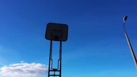 Silhouette-of-old-basketball-hoop-against-blue-sky,-low-angle-orbit-view