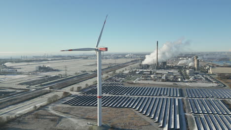 Windturbine-Dreht-Sich-In-Frostiger-Landschaft,-Rauchende-Industrieanlage,-Sonnenkollektoren,-Luftdrohnenaufnahme