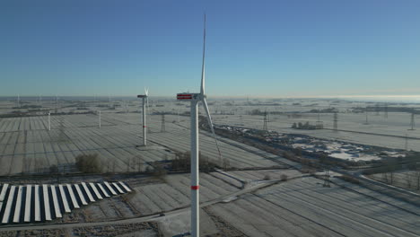 Wind-turbines-spinning-in-frosty-landscape,-bright-blue-sky,-slow-moving-aerial-drone-shot