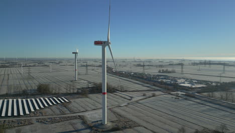Wind-turbines-spinning-in-frosty-landscape,-bright-blue-sky,-static-aerial-drone-shot