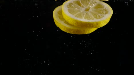 Fresh-lemon-slices-are-thrown-into-water-in-slo-mo,-black-background